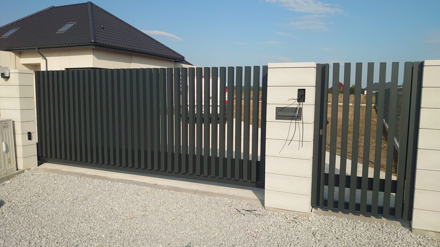 brama i furtka aluminiowa pionowa, osadzone na kamiennych białych bloczkach. W tle dach domu mieszkalnego oraz niebo