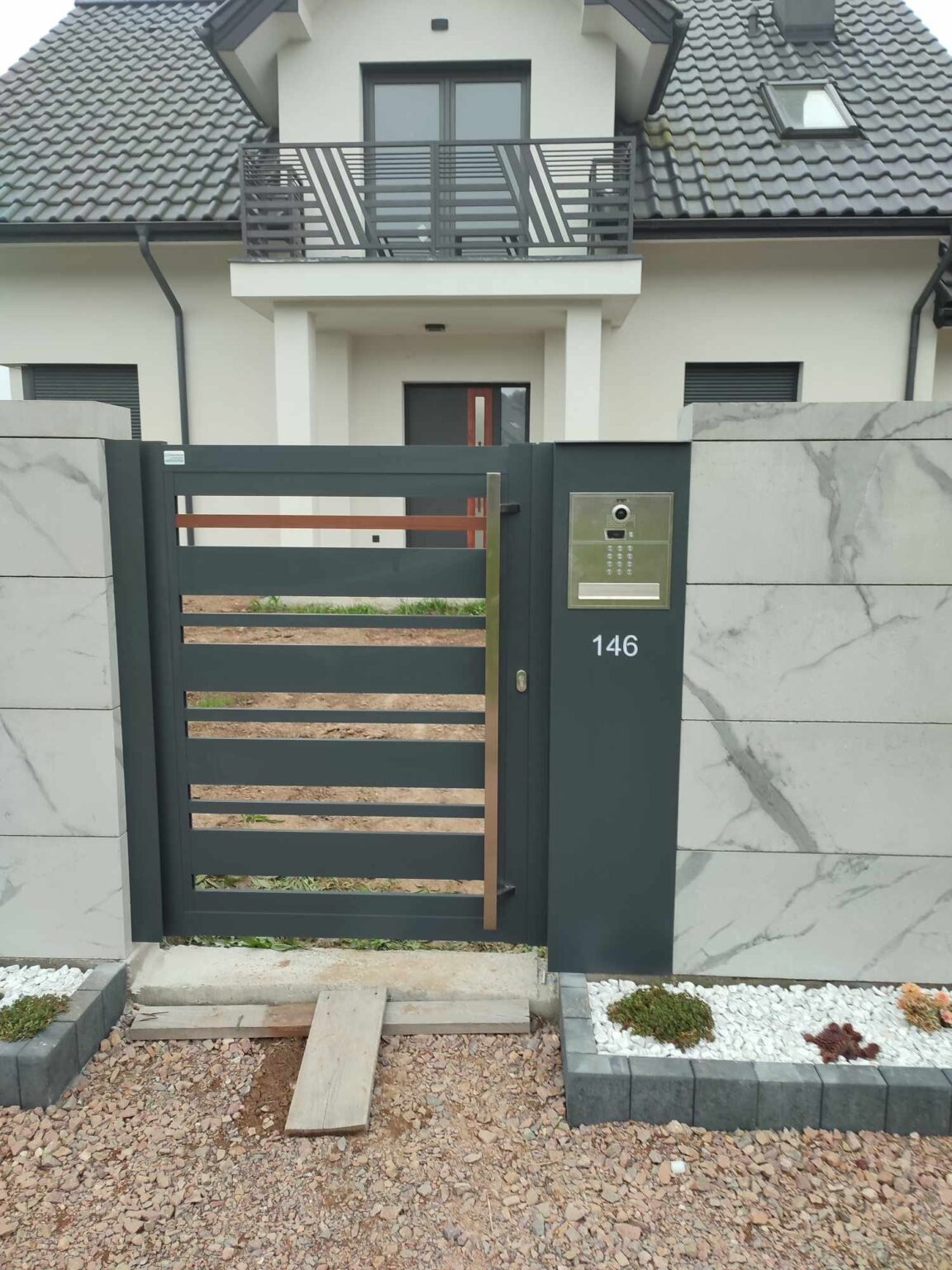 furtka aluminiowa ze słupkiem pocztowym, osadzona na kamiennych bloczkach w kolorze marmuru, w tle elewacja i wejście do budynku mieszkalnego