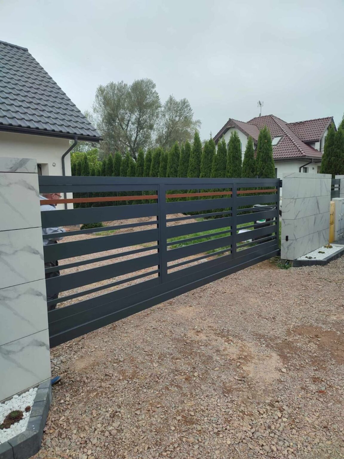 brama aluminiowa w kolorze grafitu, osadzona na kamiennych bloczkach, w tle drzewa, krzewu i dachy budynków mieszkalnych