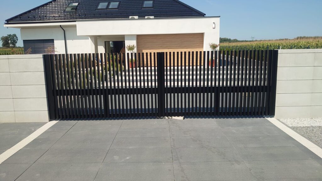 Brama aluminiowa widok z przodu. Kolor grafitowy. W tle dom z garażem i zielony krajobraz