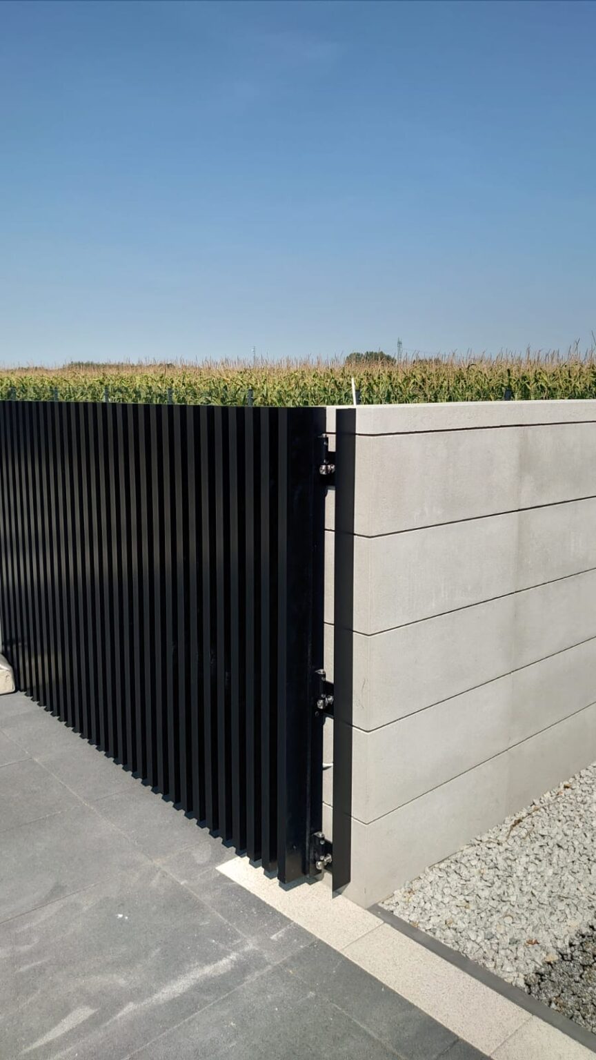 widok na ogrodzenie aluminiowe comb standard o pionowym układzie profili, kolor grafitowy, po prawej stronie bloczek ogrodzeniowy w kolorze kremowej bieli, w tle niebo i pole uprawne