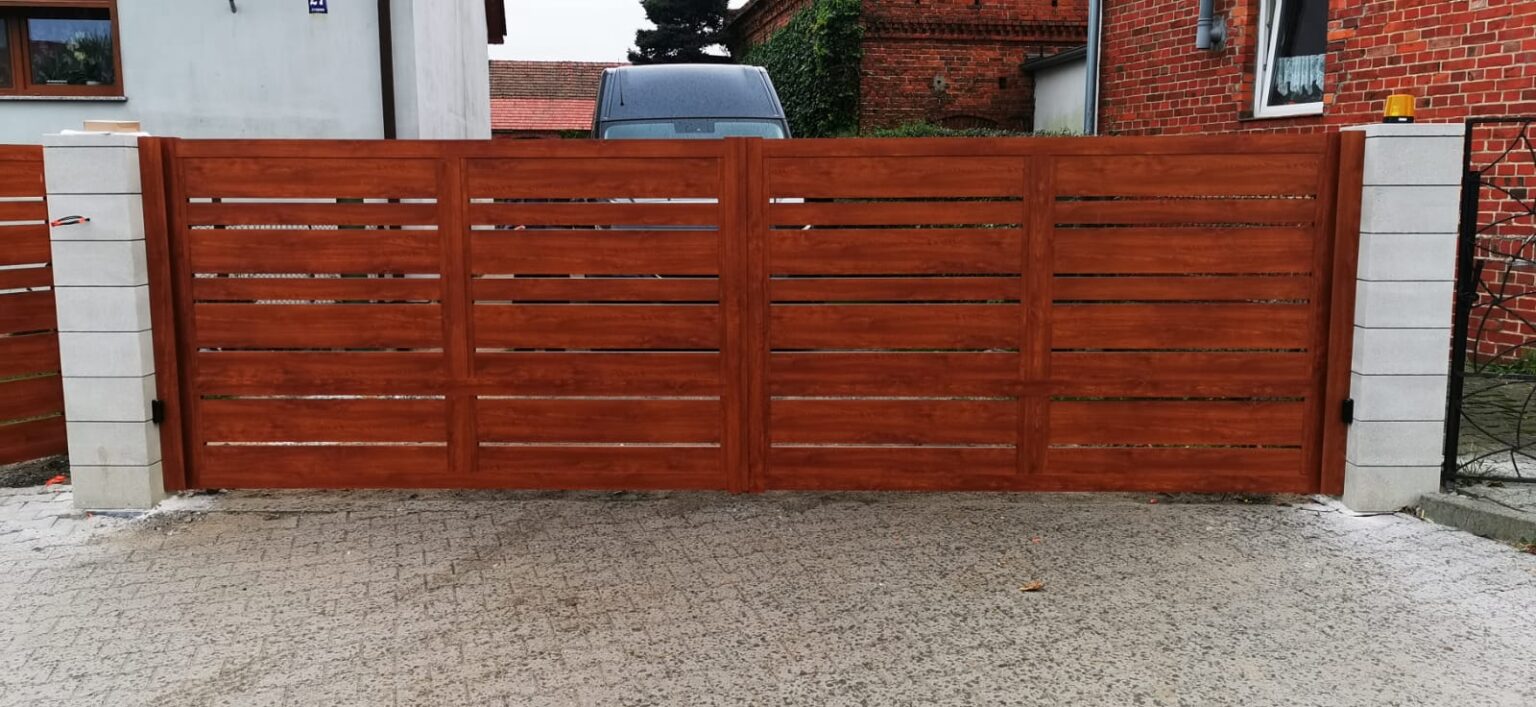 Ogrodzenie aluminiowe w kolorze termodruku drewnopodobnego, osadzone na betonowych bloczkach. Na pierwszym planie podjazd, w tle elewacja budynku, drzewa, samochód