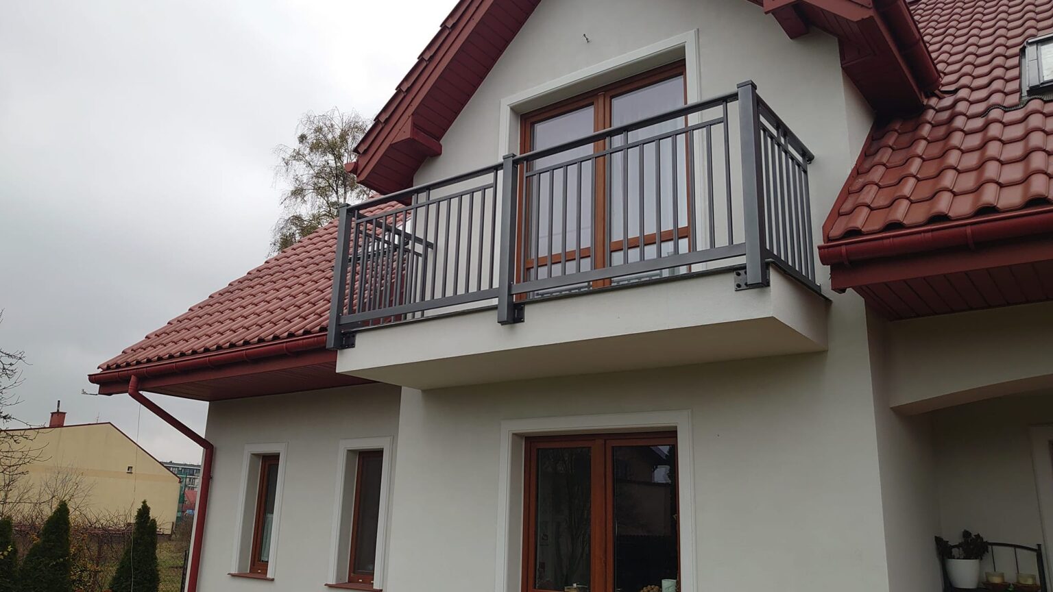 Balkonowa balustrada aluminiowa Sunny w kolorze grafitu, na drugim planie budynek i jego elewacja, w tle inne budynki oraz krzewy