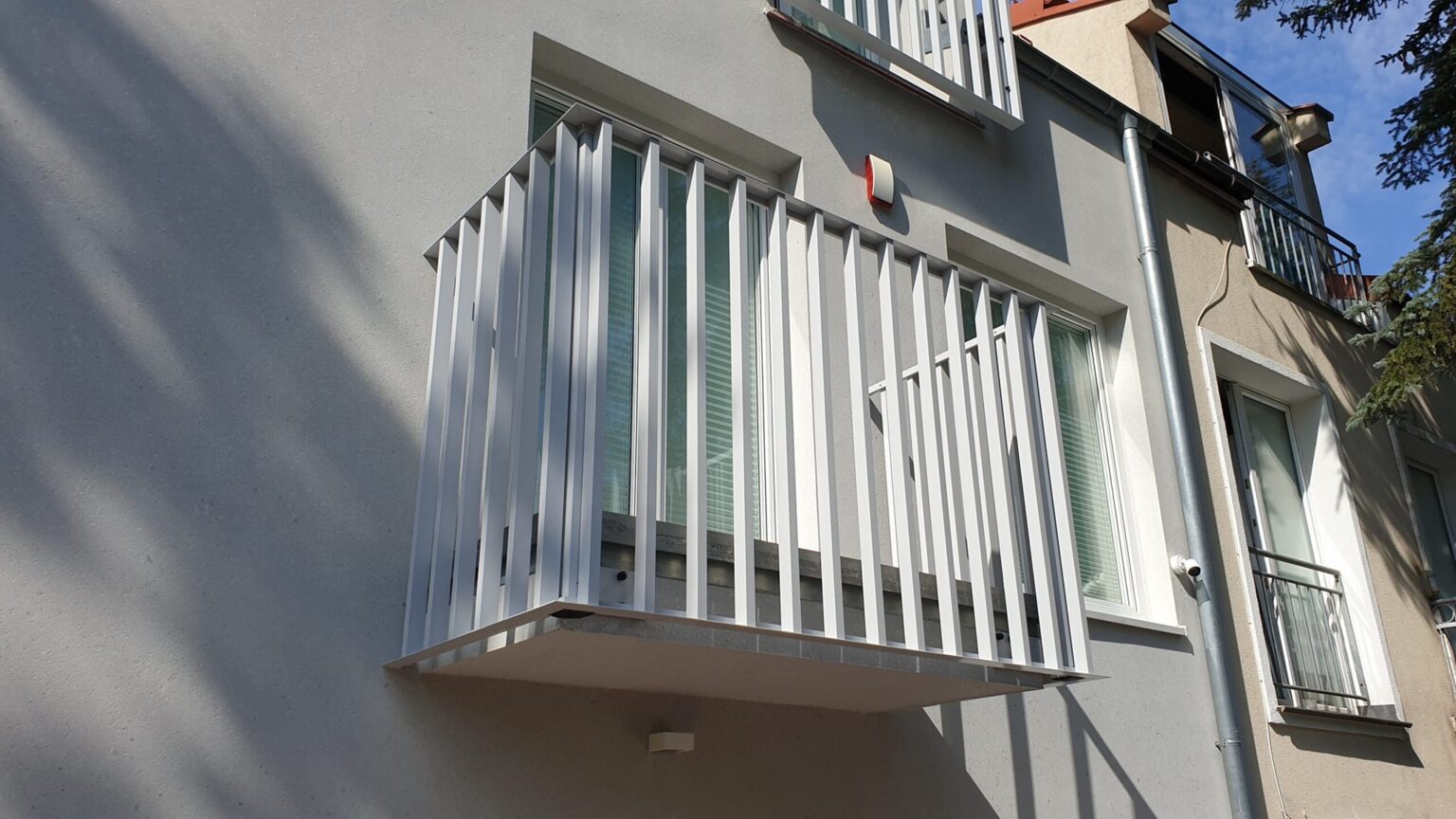 Aluminiowa balustrada comb light w kolorze białym, pionowe profile typu grzebieniowego, w tle elewacja budynku oraz okna