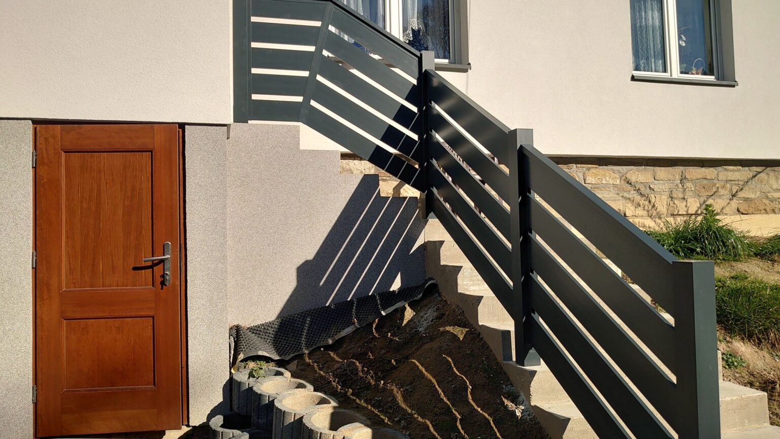 balustrada aluminiowa w kolorze grafitu, w tle elewacja budynku z oknami i boczne drzwi wejściowe