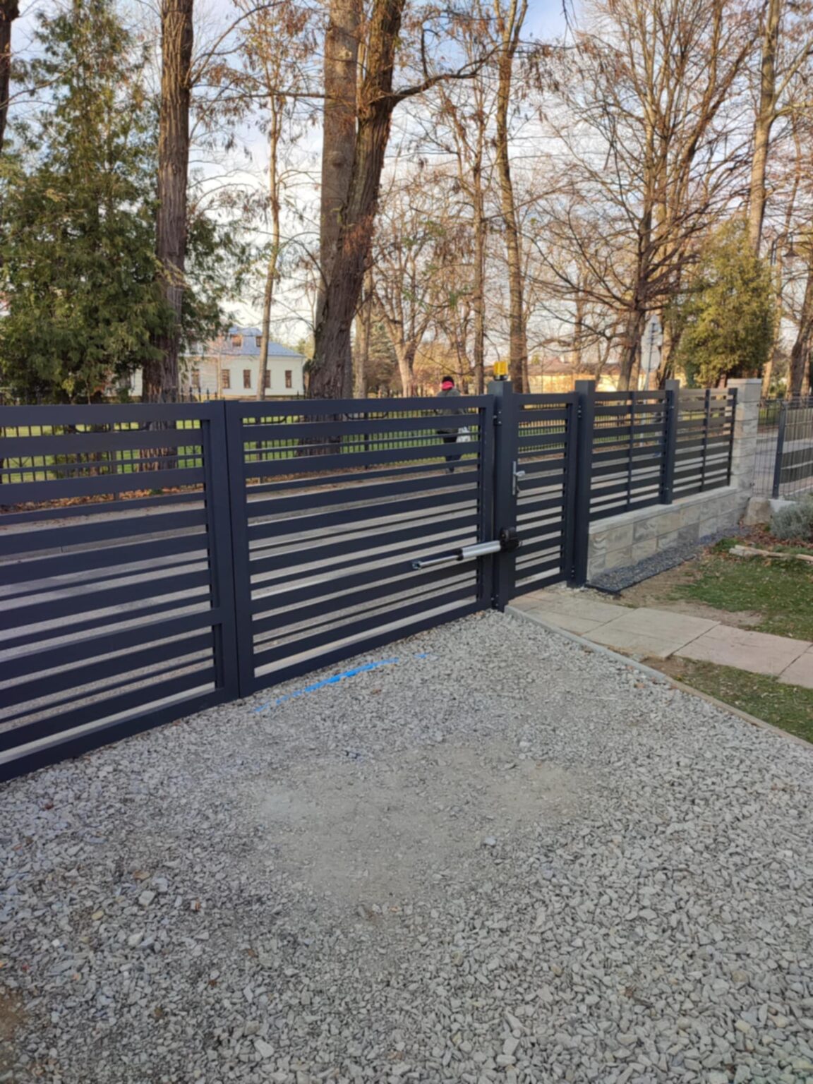 Ogrodzenie aluminiowe Friz Euro Fences, brama z frutką, w tle drzewa bez liści, na pierwszym planie ogrodzenie i kliniec