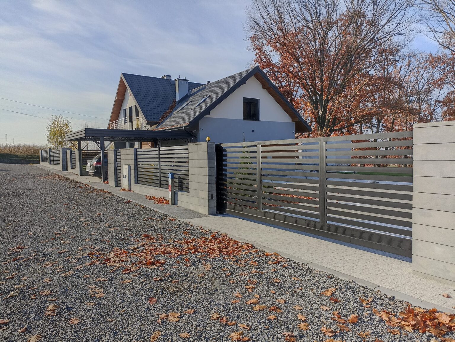 ogrodzenie aluminiowe poziome w kolorze grafitu. Na pierwszym plane podjazd z kamienia z opadniętymi jesiennymi liśćmi, w tle dom, niebo oraz drzewo