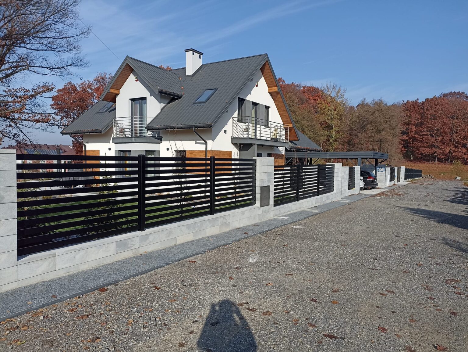 widok z bocznej perspektywy na ogrodzenie aluminiowe poziome w kolorze grafitu. Na pierwszym planie podjazd z klińca kamiennego, w tle budynek, niebo oraz wierzchołki drzew