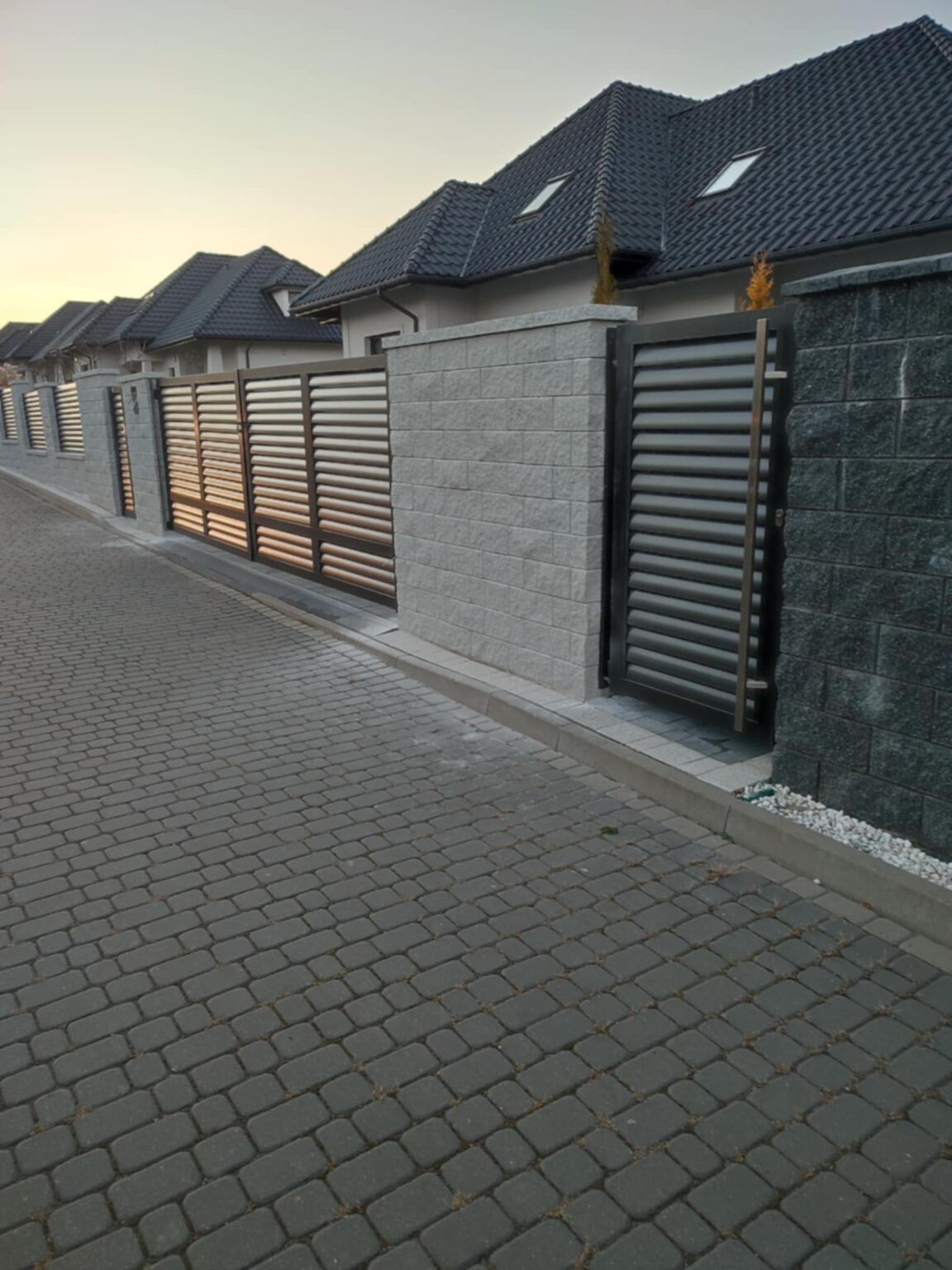 Ogrodzenie aluminiowe, furtka i brama, w tle przęsła, wszystko w kolorze grafitowym z czarnymi akcentami na ramach, umieszczone na kremowych kamiennych bloczkach ogrodzeniowych. W tle dachy i elewacje budynków, na pierwszym planie podjazd z kostki brukowej