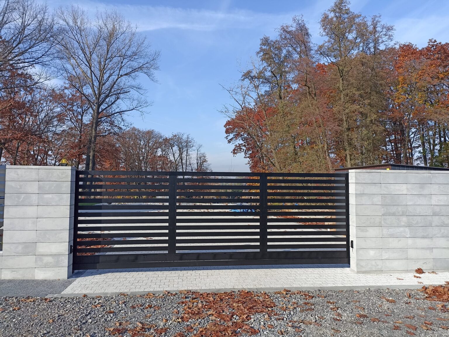 brama aluminiowa w kolorze grafitu, umieszczona na kamiennych bloczkach w kolorze kremowym. Na pierwszym planie klieniec kamienisty oraz jesienne liście, w tle niebo oraz wierzchołki drzew