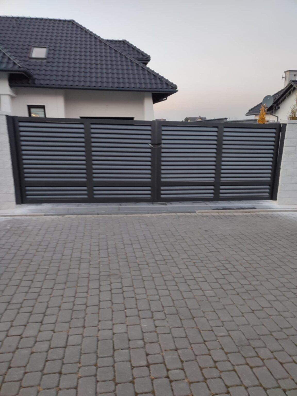 Brama aluminiowa louvre typu żaluzjowego, w kolorze grafitu, rama z akcentami czerni. Na pierwszym planie podjazd z kostki brukowej, w tle niebo, elewacja budynku i czarny dach
