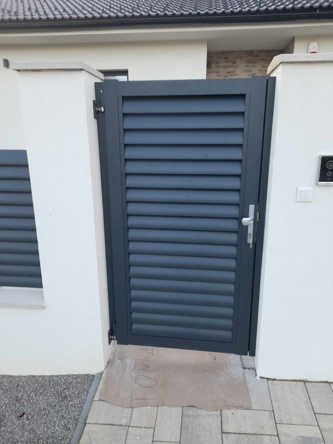 furtka aluminiowa w kolorze grafitu, osadzona na betonowych, białych bloczkach, w tle część elewacji budynku