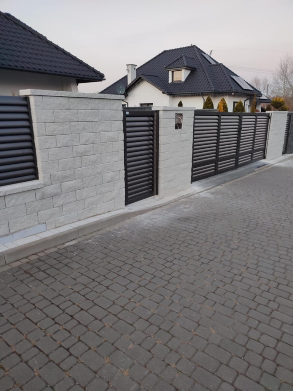 ogrodzenie aluminiowe louvre, typu żaluzjowego, umieszczone na kamiennych, kremowych bloczkach. W tle elewacje budynków i czarne dachy, na pierwszym planie podjazd z kostki brukowej